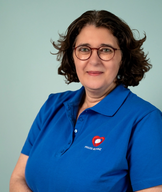 Porträt von Frau Dr. med. C. Carvalho, Ärztin der Praxis Boyraz, mit runder Brille und dunklem, lockigem Haar im blauen Praxis-Poloshirt lächelnd vor einer salbeigrünen Wand.