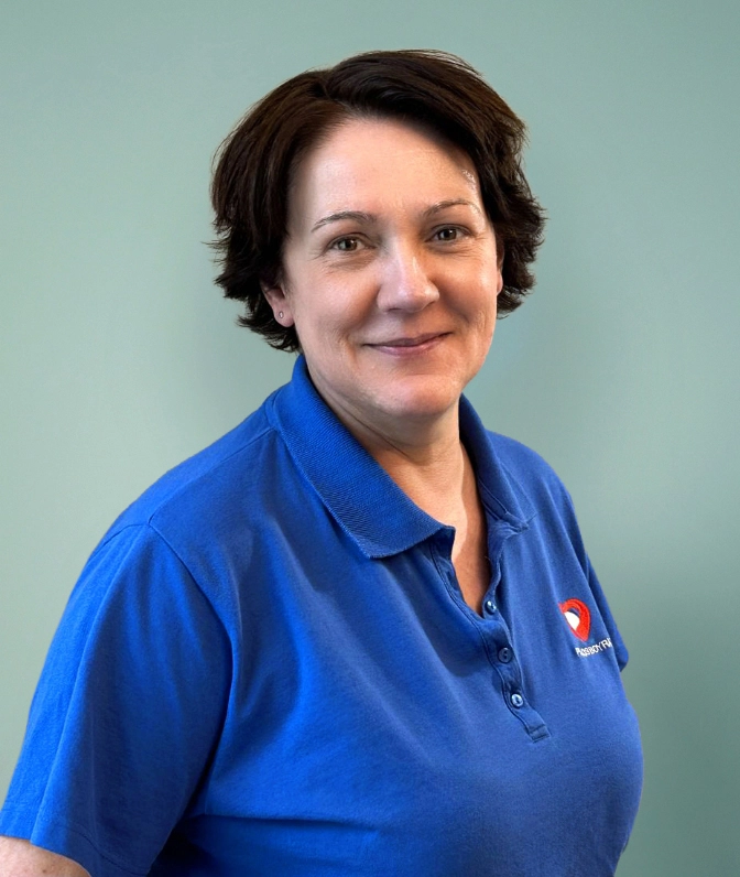 Porträt von Frau D. Devic, medizinische Fachangestellte der Praxis Boyraz, mit kurzen dunklen Haaren im blauen Praxis-Poloshirt lächelnd vor einer salbeigrünen Wand.