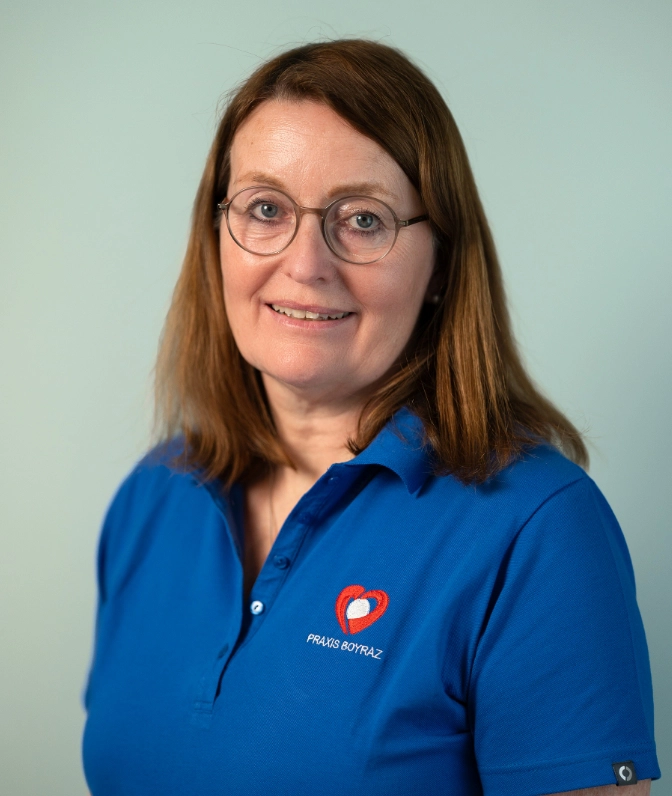 Porträt von Frau Dr. med. U. Seuss, Ärztin der Praxis Boyraz, mit Brille im blauen Praxis-Poloshirt lächelnd vor einer hellen, mintgrünen Wand.