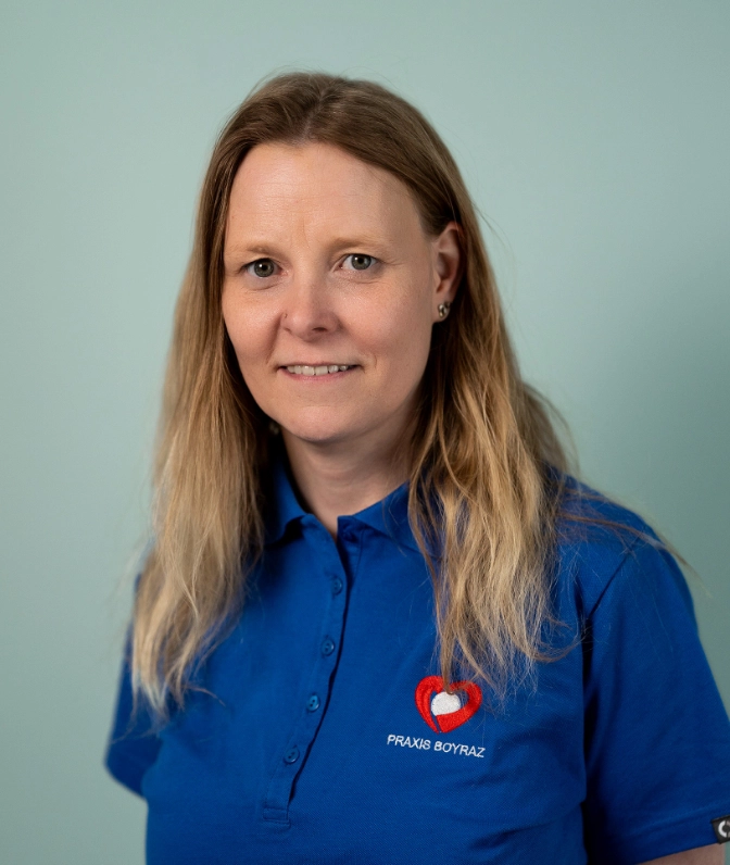 Porträt von Frau F. Böhm, Medizinische Fachangestellte der Praxis Boyraz, im blauen Praxis-Poloshirt lächelnd vor einer salbeigrünen Wand.