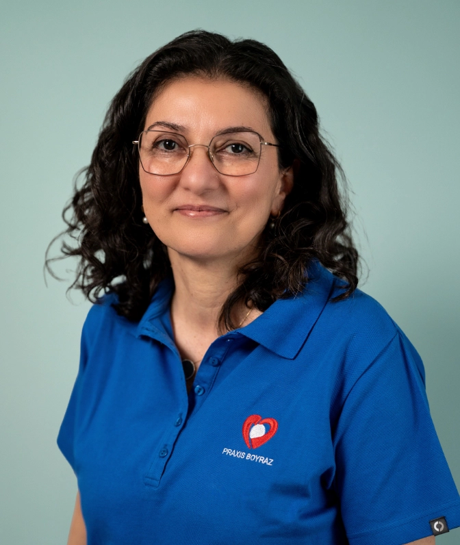 Porträt von Frau S. Boyraz, Inhaberin der Praxis Boyraz, mit Brille und dunklen, lockigen Haaren im blauen Praxis-Poloshirt lächelnd vor einer salbeigrünen Wand.