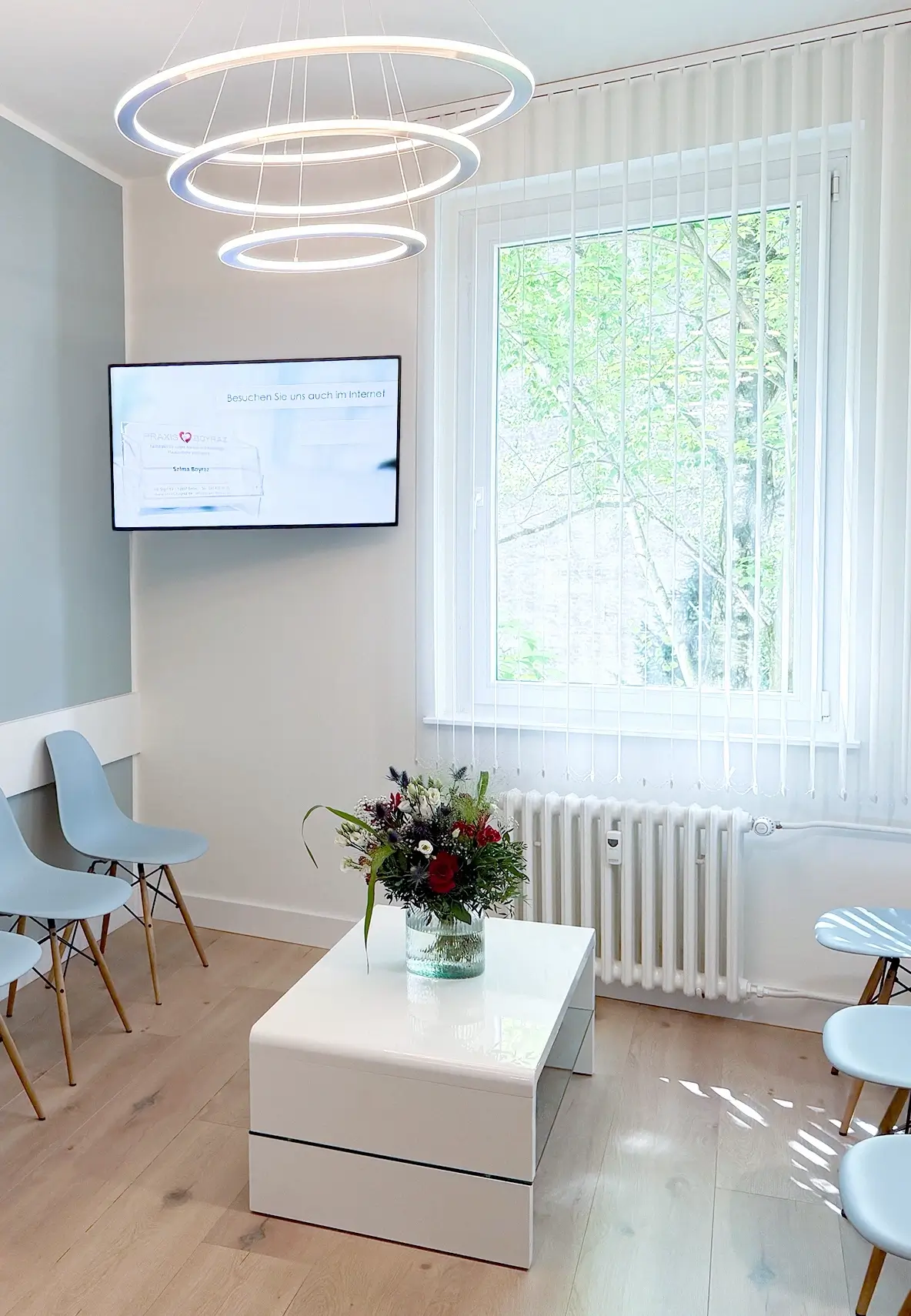 Heller Wartebereich der Praxis Boyraz mit großem Fenster, Informationsbildschirm an der Wand, einem Couchtisch mit Blumen und modernen Ring-Pendelleuchten.