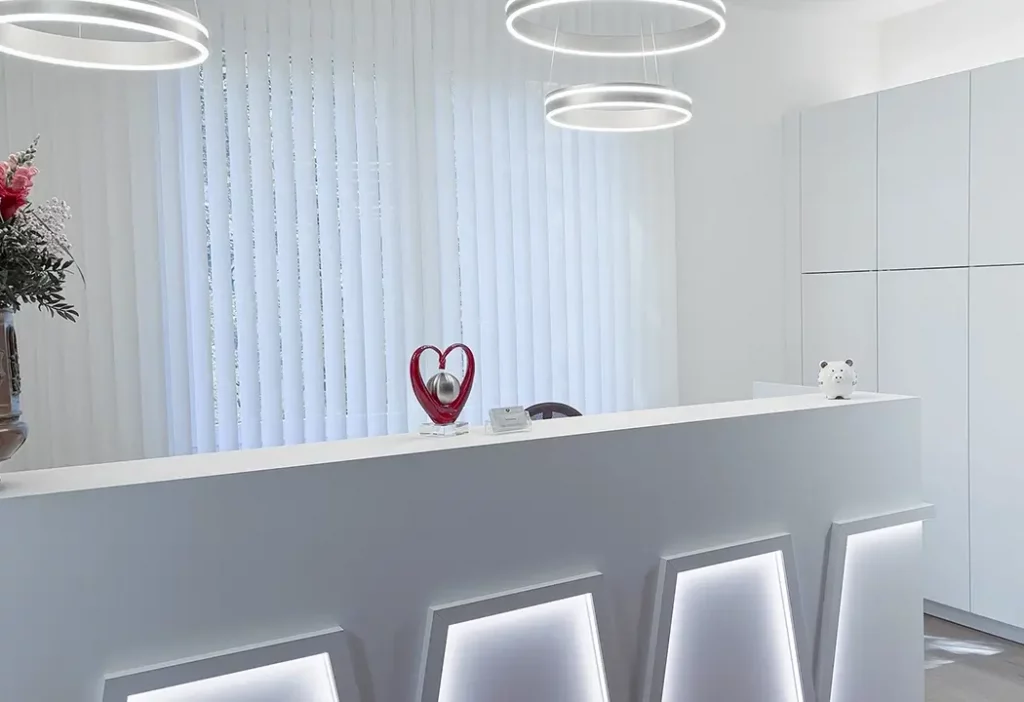 Moderner, weißer Empfangstresen der Praxis Boyraz mit LED-Beleuchtung, einer roten Herz-Skulptur und weißen Lamellen im Hintergrund.